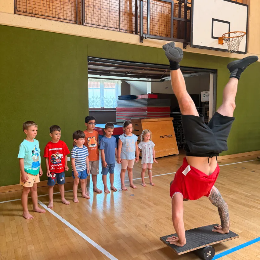 In einem Gymnastiksaal macht ein Mann in einem roten T-Shirt einen Handstand auf einem Brett, während Kinder in Shorts und T-Shirts zuschauen. Ein Basketballkorb ist an der Wand montiert.