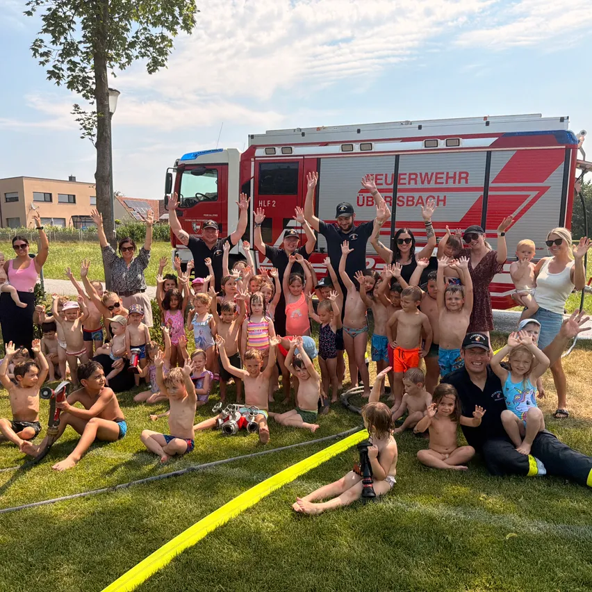 Eine Gruppe von Kindern und Erwachsenen posiert für ein Foto vor einem Feuerwehrwagen. Der Feuerwehrwagen ist in einem Grasbereich geparkt. Einige der Kinder tragen Badekleidung.