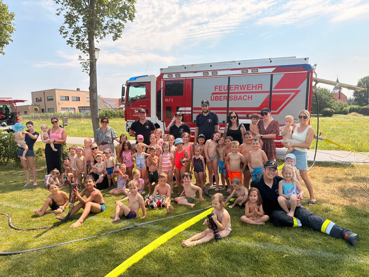 Eine Gruppe von Kindern und Erwachsenen posiert für ein Foto vor einem roten Feuerwehrauto. Einige tragen Badekleidung und Sonnenbrillen. Die Umgebung ist ein saftiger Rasen.