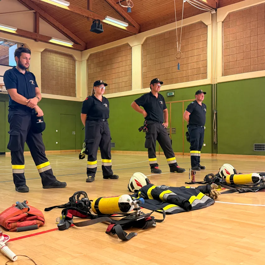 Vier Feuerwehrleute stehen in einem Raum mit Holzböden, in schwarzen Uniformen gekleidet. Helme, Sauerstoffflaschen und andere Ausrüstung sind verstreut.