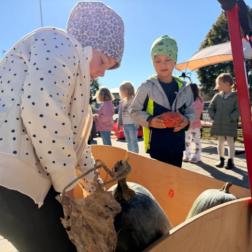 Ein junges Mädchen mit einem Leopardenmuster-Kopftuch pflückt Kürbisse aus einem Holzwagen. Ein Junge mit grüner Mütze hält einen Kürbis in den Händen.