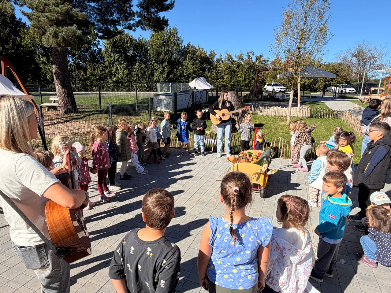 Eine Frau spielt Gitarre, während eine Gruppe von Kindern zuschaut. Ein Mann steht neben ihr und spielt ebenfalls Gitarre.