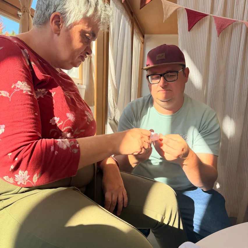 Ein Mann und eine Frau sitzen zusammen. Der Mann trägt eine Mütze und eine Brille und hält ein kleines Objekt. Die Frau trägt ein Blumenhemd und schaut den Mann an.