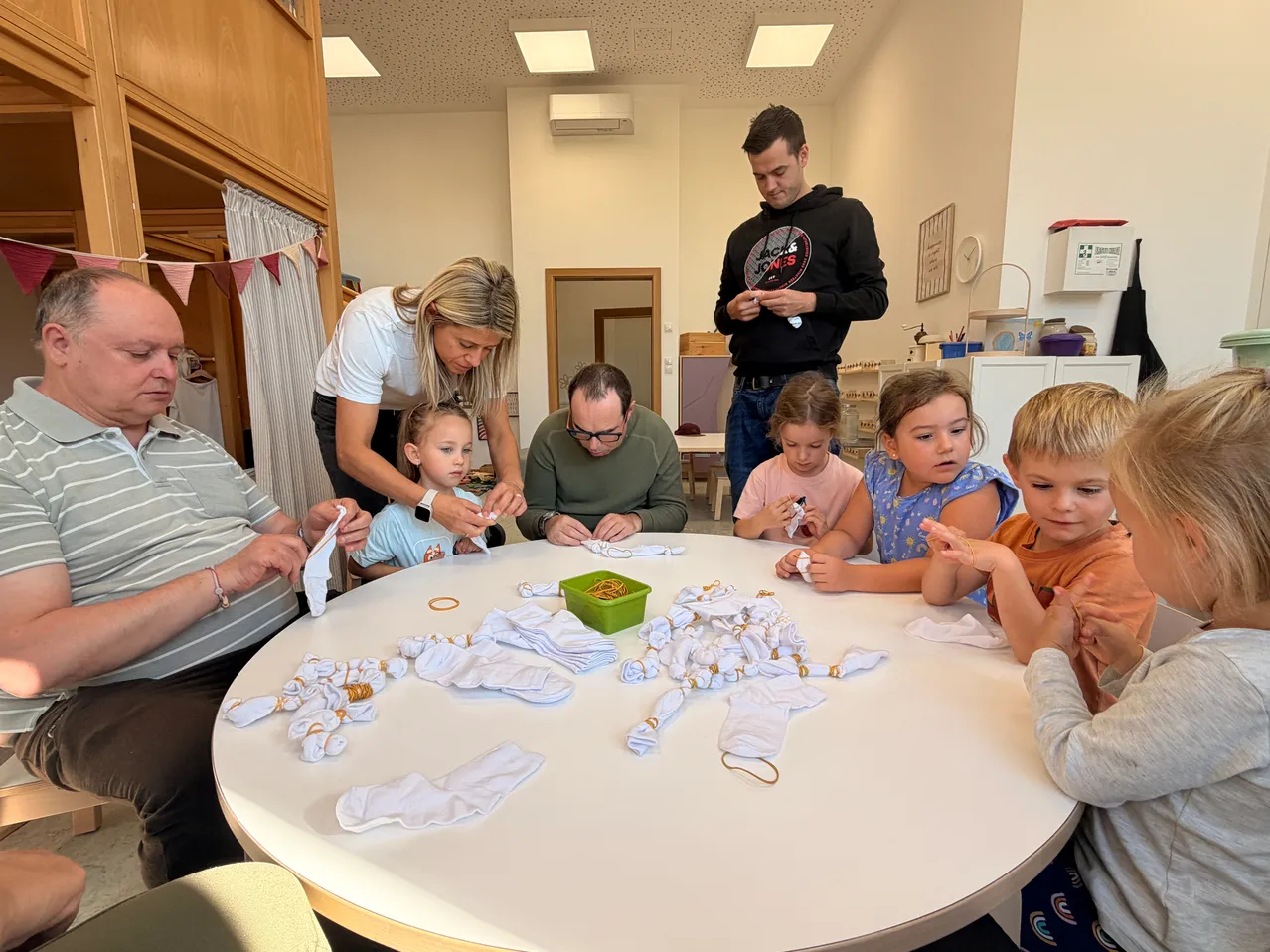Eine Gruppe von Kindern und Erwachsenen ist um einen Tisch versammelt und beschäftigt sich mit einer Bastelaktivität. Der Tisch ist mit weißem Tuch bedeckt und hat verschiedene Materialien darauf, einschließlich Socken und anderer Gegenstände. Die Kinder tragen Freizeitkleidung und scheinen sich auf ihre Aktivität zu konzentrieren.