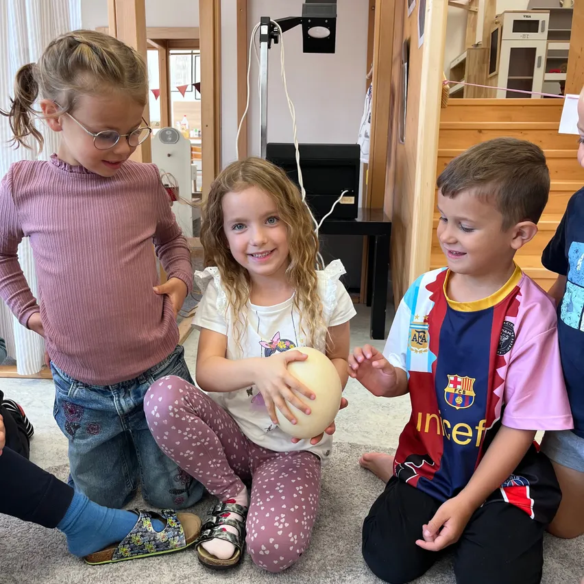 Drei Kinder sitzen auf dem Boden, eines hält ein Ei. Das Mädchen in der Mitte lächelt und hält ein Ei. Der Junge rechts trägt ein Barcelona-Fußballtrikot.
