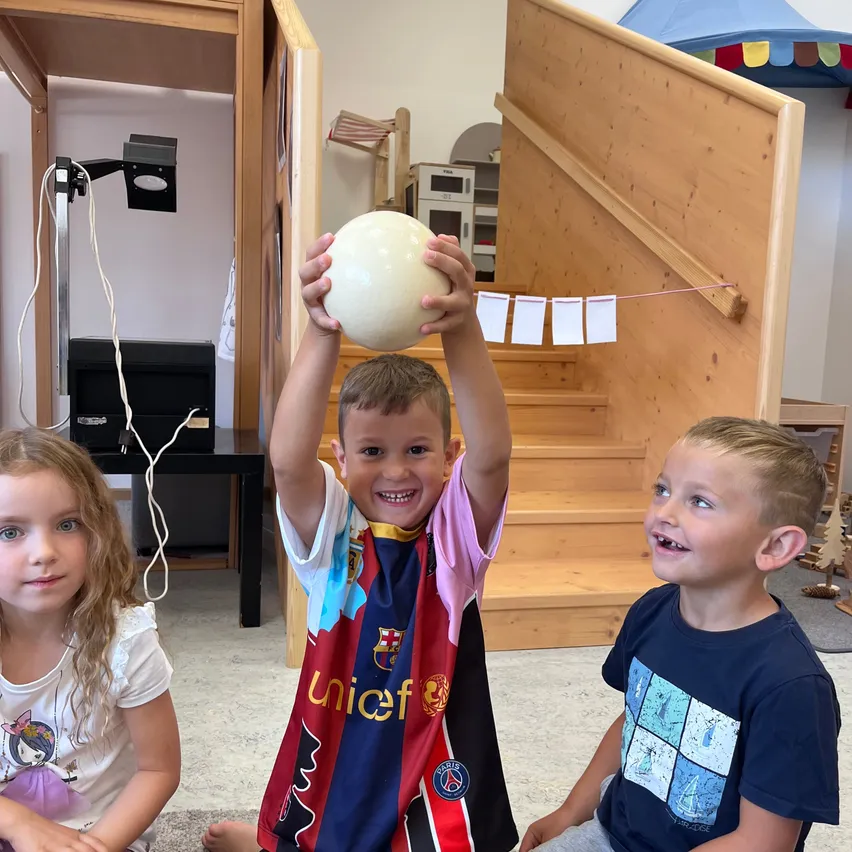 Drei Kinder sitzen auf dem Boden in einem Raum. Ein Junge in der Mitte hält einen großen Ball und lächelt. Das Mädchen auf der linken Seite schaut den Jungen an. Der Junge auf der rechten Seite lächelt und schaut in die Kamera.