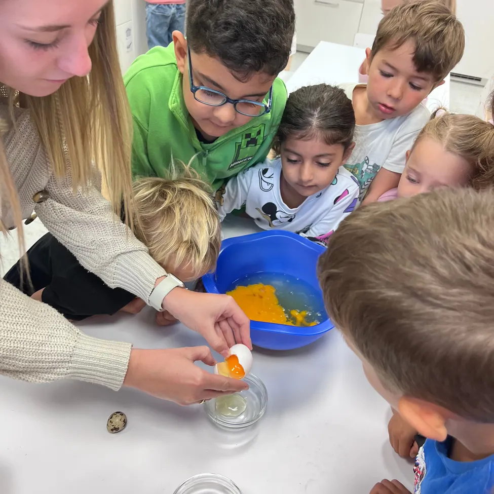 Eine Frau und mehrere Kinder sind um einen Tisch versammelt, wo die Frau ein Ei in eine Schüssel bricht.