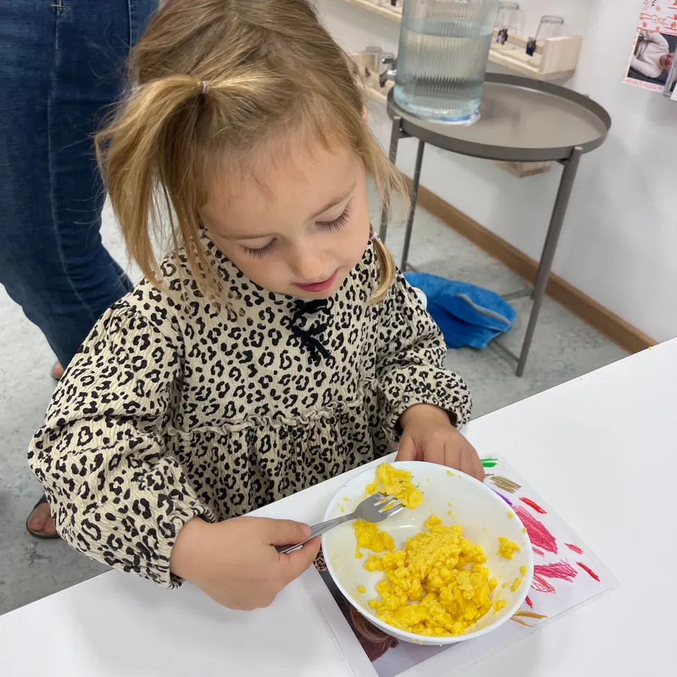 Ein kleines Mädchen mit blonden Haaren sitzt an einem Tisch und isst eine Schüssel gelbes Essen mit einer Gabel.