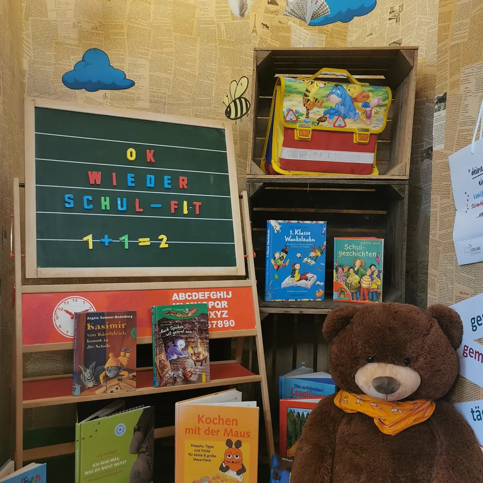 Ein Klassenzimmer-Setup mit einem Tafelschild, das 'OK WIEDER SCHUL-FIT 1+1=2' sagt. Ein brauner Teddybär mit einem Schal sitzt neben Büchern und einer Schultasche.