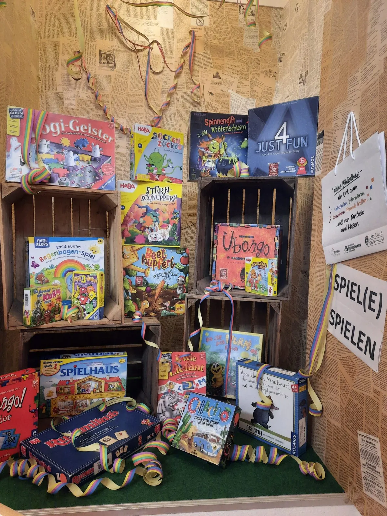 Eine Ausstellung verschiedener Brettspiele auf Holzregalen mit bunten Dekorationen, darunter 'Oggy und die Kakerlaken', 'Socken Zocken' und 'Spinnendif und Kürotenschleim'. Ein weißer Beutel hängt an der Wand mit einem Schild, das 'SPIEL(E) SPIELEN' sagt.