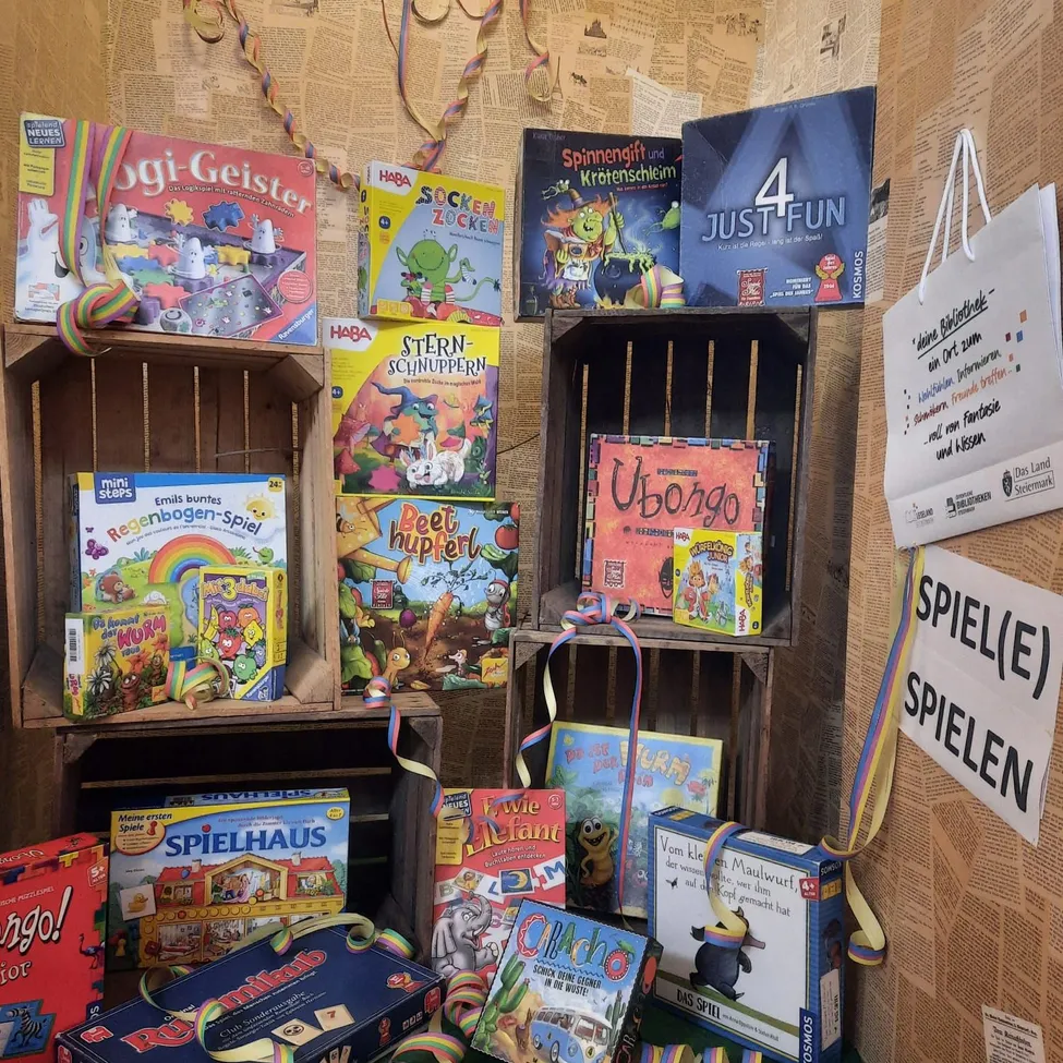 Eine Ausstellung verschiedener Brettspiele auf Holzregalen mit bunten Dekorationen, darunter 'Oggy und die Kakerlaken', 'Socken Zocken' und 'Spinnendif und Kürotenschleim'. Ein weißer Beutel hängt an der Wand mit einem Schild, das 'SPIEL(E) SPIELEN' sagt.