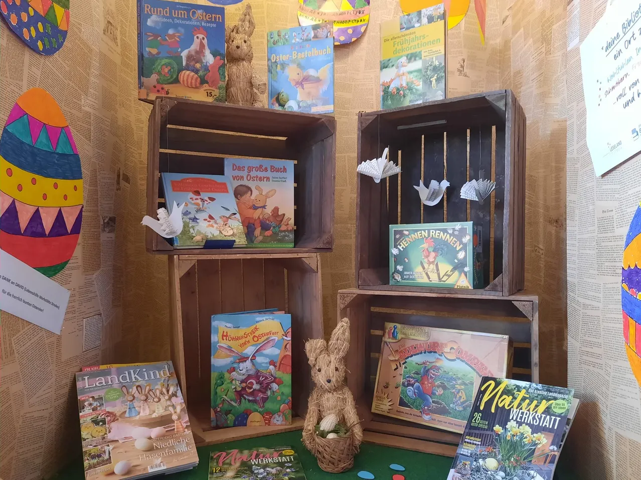 Verschiedene Osterbücher und Dekorationen sind auf Holzkisten angeordnet. Eine Hasenfigur steht auf einem grünen Teppich. Der Hintergrund besteht aus Zeitungsausschnitten.