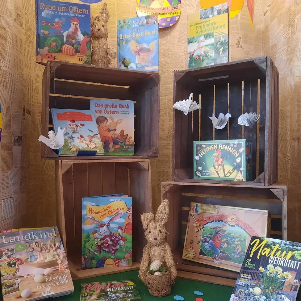 Verschiedene Osterbücher und Dekorationen sind auf Holzkisten angeordnet. Eine Hasenfigur steht auf einem grünen Teppich. Der Hintergrund besteht aus Zeitungsausschnitten.