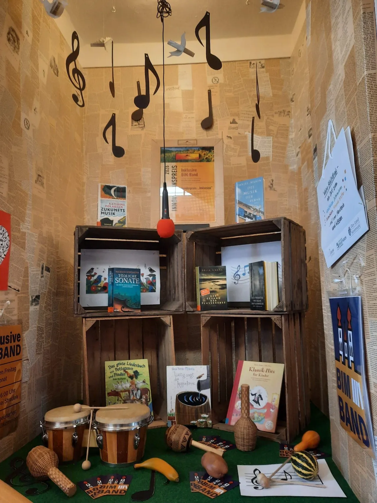 Eine Ecke mit Holzkisten gefüllt mit Büchern und Musikinstrumenten. Noten sind an der Wand und ein roter Luftballon hängt.