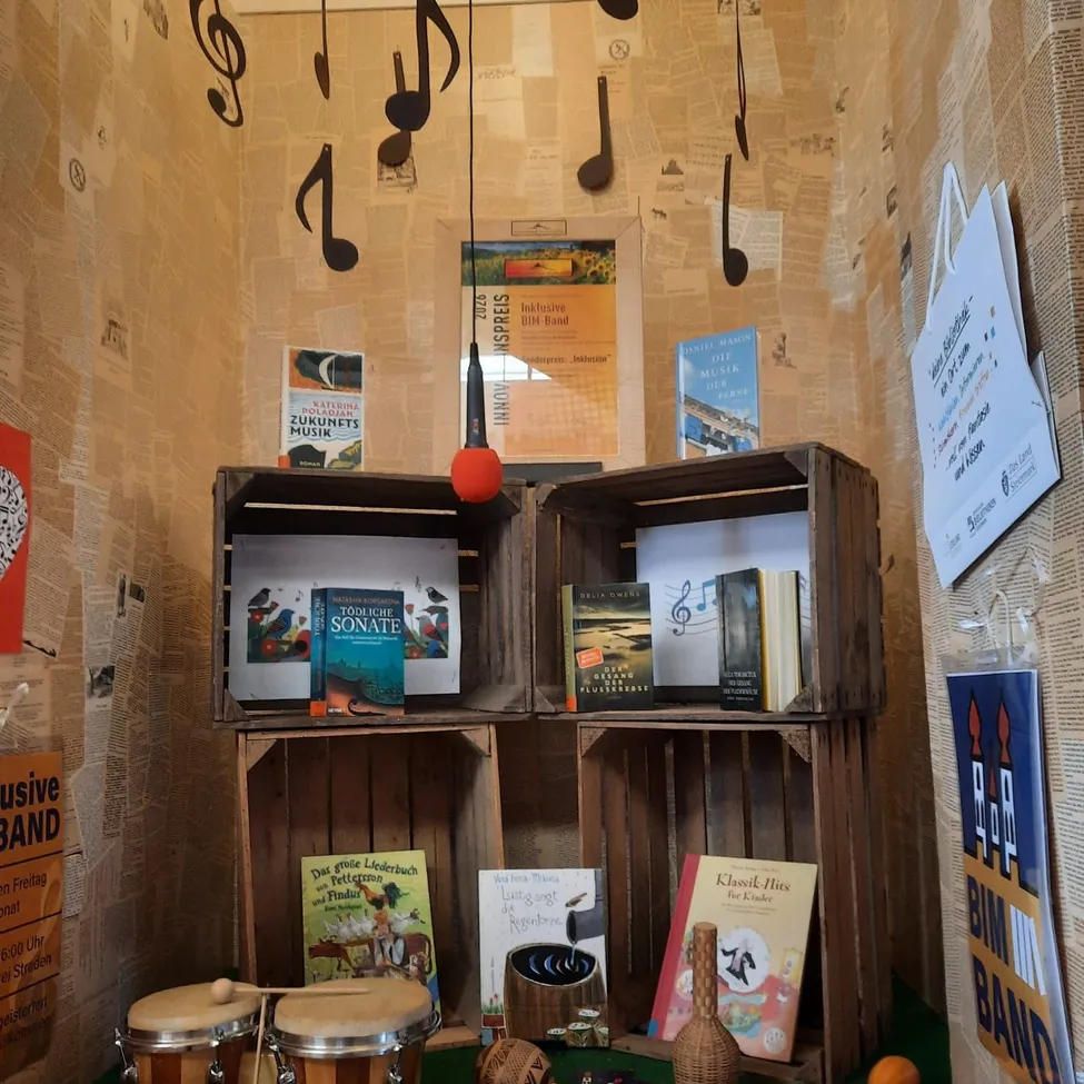 Eine Ecke mit Holzkisten gefüllt mit Büchern und Musikinstrumenten. Noten sind an der Wand und ein roter Luftballon hängt.