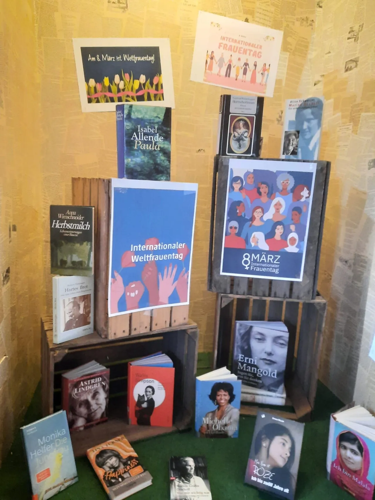 Eine Ausstellung von Büchern mit einem Plakat, das Internationaler Frauentag darauf steht. Die Bücher sind in Kisten und auf Regalen angeordnet.