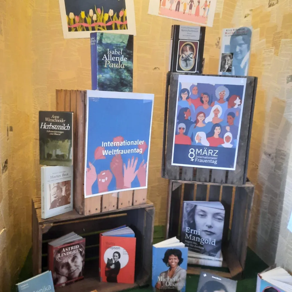 Eine Ausstellung von Büchern mit einem Plakat, das Internationaler Frauentag darauf steht. Die Bücher sind in Kisten und auf Regalen angeordnet.