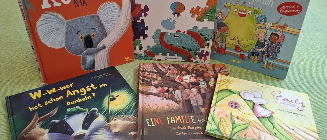 Eine Sammlung von Kinderbüchern auf einer grünen Oberfläche. Die Bücher umfassen 'Trau Dich Koala Bar', 'Einzigartig', 'Als Kleine Wie Im Kindergarten', 'W-W-Wer Hat Schon Angst Im Dunkeln?' und 'Eine Familie Wie Zuckerbrot'.