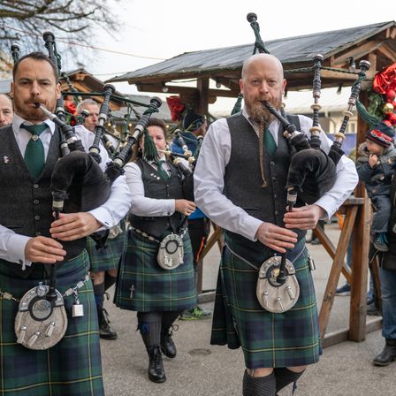 Drei Dudelsackspieler treten im Freien auf. Sie tragen Kilts und Westen. Hinter ihnen steht eine hölzerne Struktur mit Dach und Weihnachtsdekorationen. Ein Kind steht rechts und beobachtet sie.
