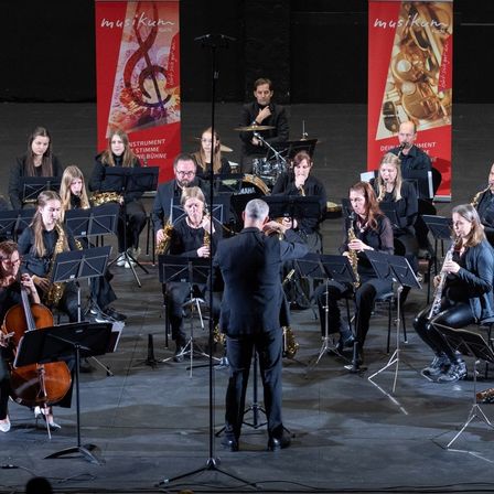 Eine Gruppe von Musikern auf der Bühne, die mit verschiedenen Instrumenten spielen. Ein Dirigent steht davor und leitet das Orchester. Im Hintergrund befinden sich Banner mit Musiknoten.