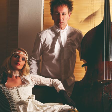 Eine Frau in einem weißen Kleid sitzt mit einem Akkordeon, während ein Mann in einem weißen Hemd hinter ihr steht, beide neben einem großen Cello.