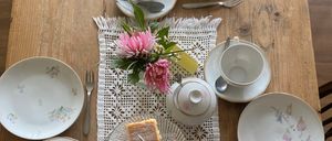 Ein hölzerner Tisch für eine Mahlzeit vorbereitet mit verschiedenen Tellern, Tassen, Untertassen, Gabeln und einer Teekanne. Es gibt Kuchenstücke auf einem Glasteller und eine Blumenarrangement.
