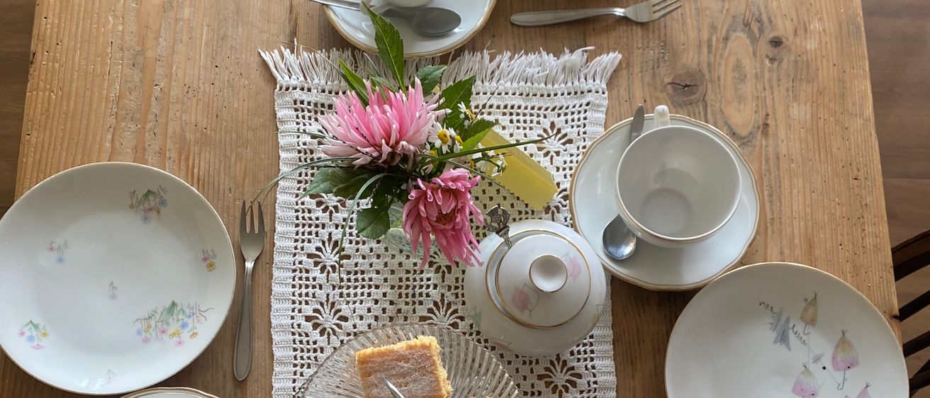 Ein hölzerner Tisch für eine Mahlzeit vorbereitet mit verschiedenen Tellern, Tassen, Untertassen, Gabeln und einer Teekanne. Es gibt Kuchenstücke auf einem Glasteller und eine Blumenarrangement.