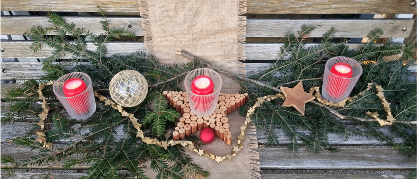 Eine Holzbank ist mit einem Jutetuch, einer Tannengirlande, einem Stern und drei roten Kerzen geschmückt. Die Kerzen stehen in Glasbechern.