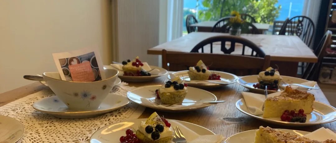 Ein Tisch mit verschiedenen Desserts wie Puddings, Kuchen und Obsttorten. Der Tisch ist aus Holz, mit Spitzendeckchen verziert und bietet eine malerische Aussicht.