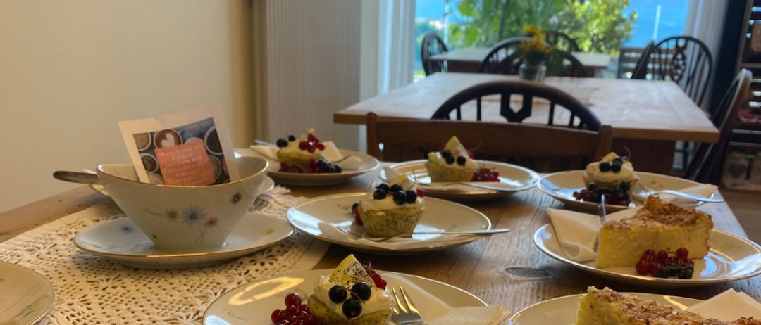 Ein Tisch mit verschiedenen Desserts wie Puddings, Kuchen und Obsttorten. Der Tisch ist aus Holz, mit Spitzendeckchen verziert und bietet eine malerische Aussicht.