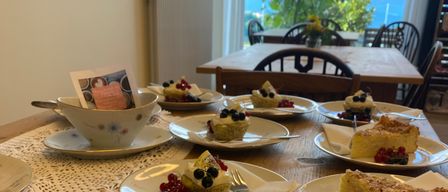 A table set with various desserts, including custards, cakes, and fruit tarts. The table is wooden, adorned with lace doilies, and overlooks a scenic view.
