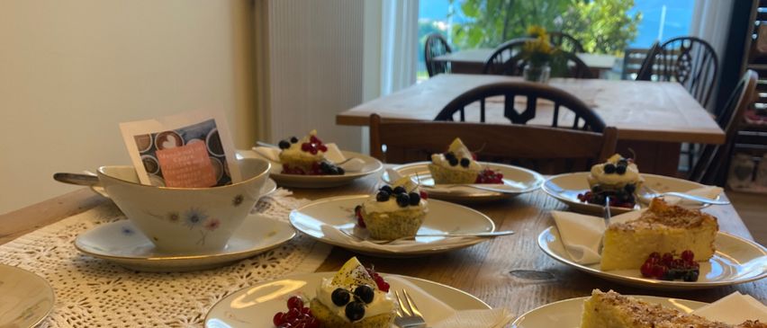 Ein Tisch mit verschiedenen Desserts wie Puddings, Kuchen und Obsttorten. Der Tisch ist aus Holz, mit Spitzendeckchen verziert und bietet eine malerische Aussicht.