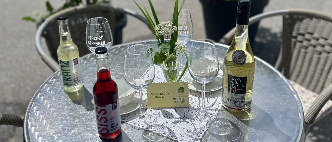 Ein kleiner Tisch mit einer Flasche Wein, einer Flasche Limonade und zwei Weingläsern. Der Tisch hat eine weiße Spitze. Es gibt zwei Topfpflanzen an der Seite.