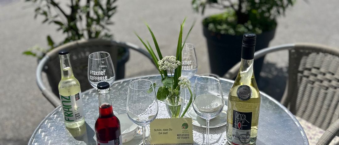 Ein runder Metalltisch mit Flaschen und Gläsern darauf. Hinter dem Tisch stehen zwei Stühle und zwei Topfpflanzen. Es gibt einen gelben Aufkleber mit Text.