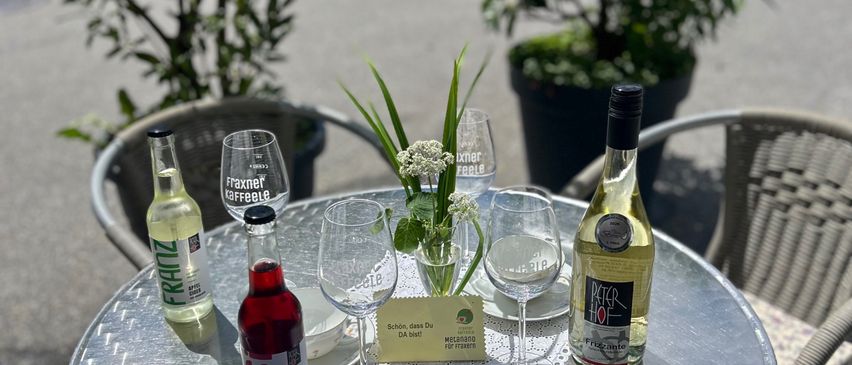 Ein runder Metalltisch mit Flaschen und Gläsern darauf. Hinter dem Tisch stehen zwei Stühle und zwei Topfpflanzen. Es gibt einen gelben Aufkleber mit Text.