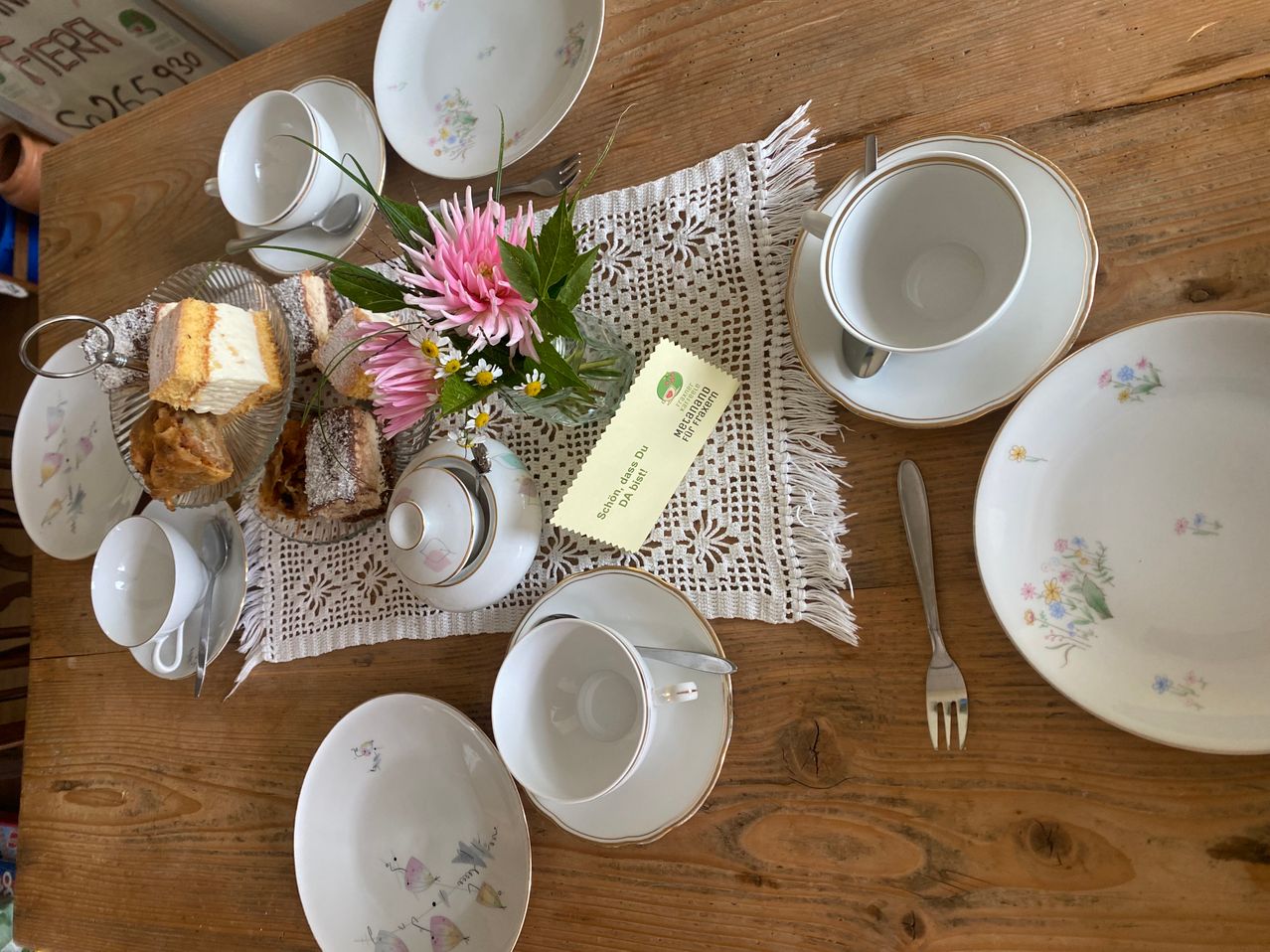 Ein hölzerner Tisch mit blumengemusterten Tellern, Tassen und Untertassen, einem Teekessel und einer Blumenarrangement. Ein gelbes Schild mit der Aufschrift 'Schokolade und Marmelade'.
