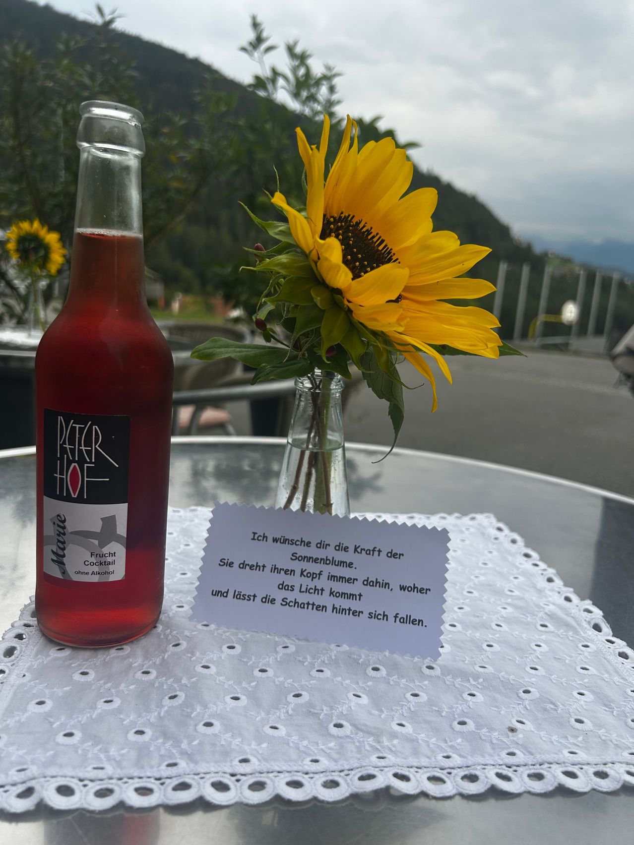 Eine Flasche mit roter Flüssigkeit mit dem Etikett 'Peter Hof' neben einer Sonnenblume in einer Vase auf einem Tisch mit einer Tischdecke und einer kleinen Karte mit einem Zitat.
