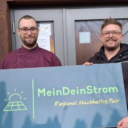 Zwei Männer halten ein Schild für MeinDeinStrom, eine regionale Nachhaltigkeitsinitiative. Sie stehen vor einer Tür mit einem Solarpanel-Grafik.