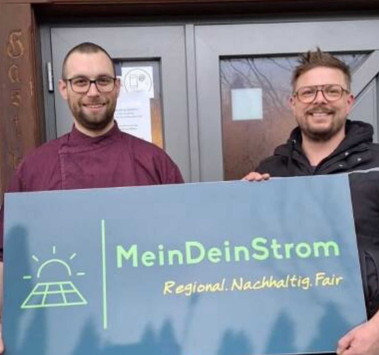 Zwei Männer halten ein Schild für MeinDeinStrom, eine regionale Nachhaltigkeitsinitiative. Sie stehen vor einer Tür mit einem Solarpanel-Grafik.
