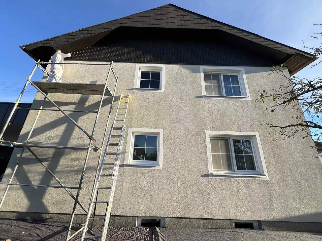 Ein Arbeiter steht auf einem Gerüst und malt ein Gebäude. Das Gebäude hat ein Ziegeldach, drei Fenster und eine Leiter an der Seite. Der Himmel ist klar und blau.