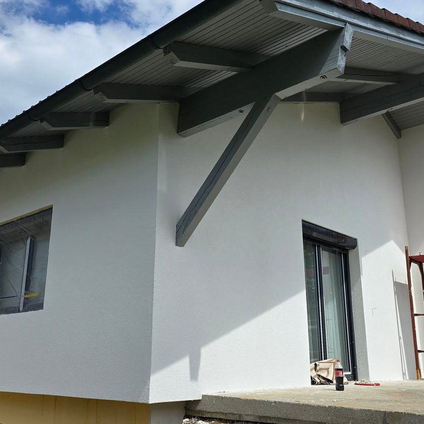 Ein weißes Haus im Bauzustand, mit einer Glastür, einem Fenster mit Schutzfolie und einer an der Wand gelehnten Leiter. Das Dach hat Dachrinnen.
