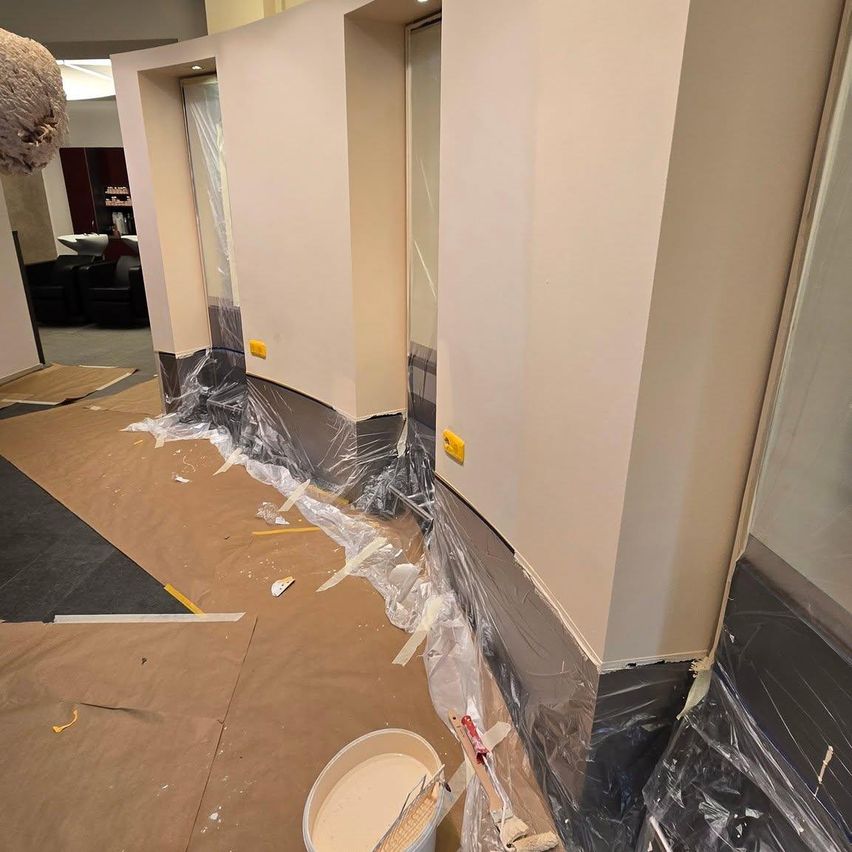 Ein Raum im Innenbereich wird renoviert, wobei Wände mit Plastik bedeckt sind, ein Eimer Farbe und Werkzeuge auf dem Boden. Zwei Türen sind teilweise bedeckt und es gibt verstreute Gegenstände auf dem Boden.