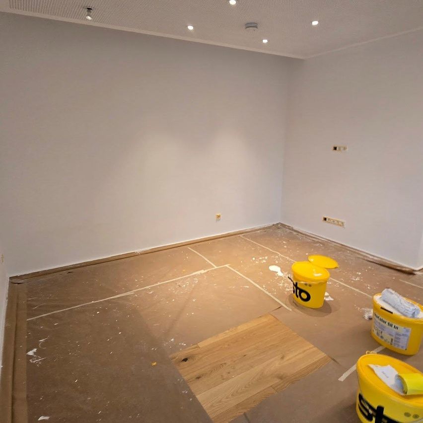 Ein leerer Raum mit weißen Wänden und einem Holzboden. Es gibt gelbe Eimer mit Farbe und Schutzpapier, das den Boden bedeckt.