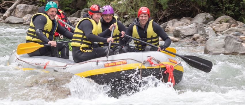 Fünf Personen in Sicherheitswesten und Helmen befinden sich in einem gelben und schwarzen Schlauchboot. Sie paddeln auf einem Fluss mit Felsen.