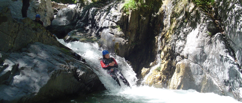 Eine Person mit blauem Helm rutscht einen Wasserfall hinunter. Zwei andere stehen auf einem Felsen darüber.