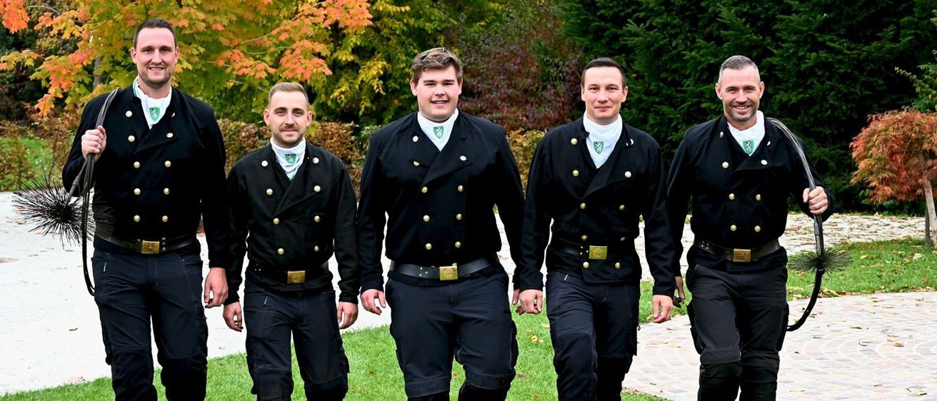 Fünf Männer in schwarzen Uniformen mit goldenen Knöpfen und Abzeichen gehen lächelnd auf einem Rasen. Hinter ihnen stehen Bäume mit Herbstlaub und ein gepflasterter Weg.