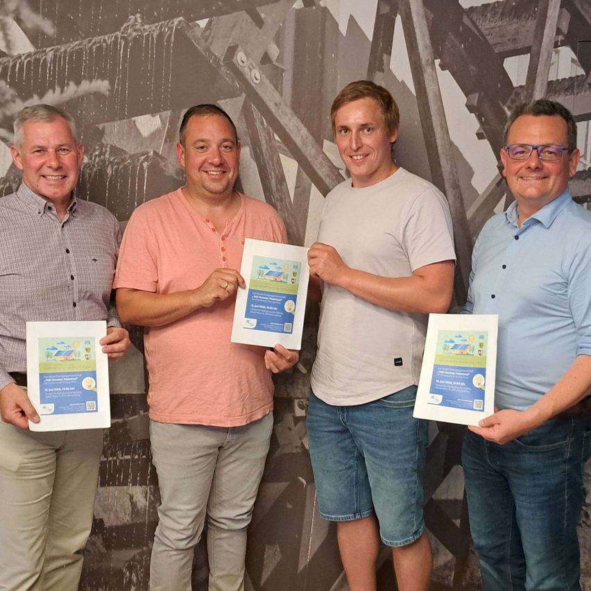 Vier Männer stehen vor einer Wandgestaltung, jeder hält ein Zertifikat in der Hand. Der Mann auf der linken Seite trägt ein kariertes Hemd, während der Mann in der Mitte ein rosa Hemd trägt. Der Mann auf der rechten Seite trägt ein blaues Hemd und eine Brille.