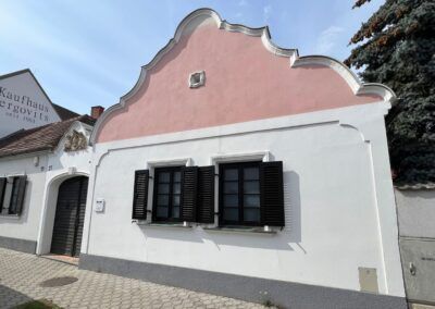 Ein weißes und rosa Gebäude mit schwarzen Fensterläden und einem Schornstein steht unter einem klaren Himmel. Das Gebäude hat einen Vintage-Stil mit Backsteinelementen und einem Schild, das 'Kaufhaim Ergovitz' steht.