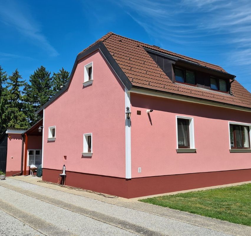 Ein rosafarbenes Haus mit braunem Dach und weißen Verkleidungen, drei Fenster im Erdgeschoss und eines im Dachgeschoss, unter einem blauen Himmel mit einigen Wolken.