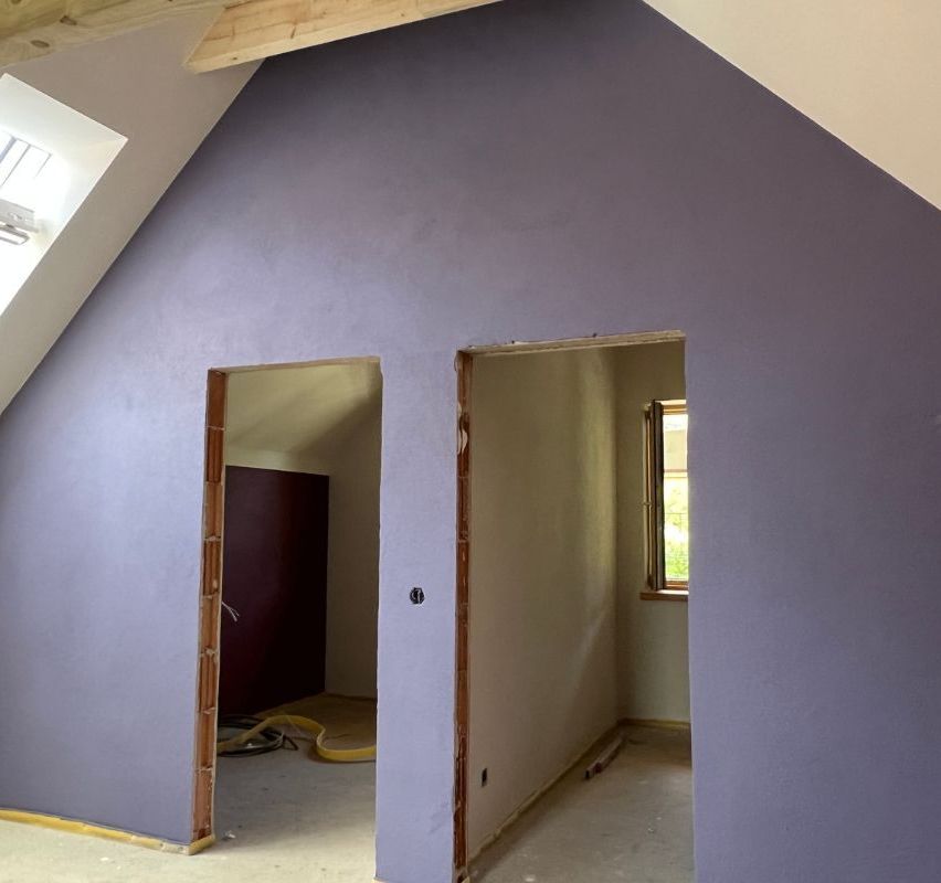 Ein Dachbodenzimmer mit lavendelfarbenen Wänden und zwei offenen Türen. Der Raum ist im Bauzustand mit freiliegenden Holzbalken und unfertigen Wänden. Ein Fenster lässt natürliches Licht herein.